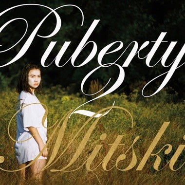 Young woman standing in a grassy field with large stylized text reading Puberty Mitski overlaying the image